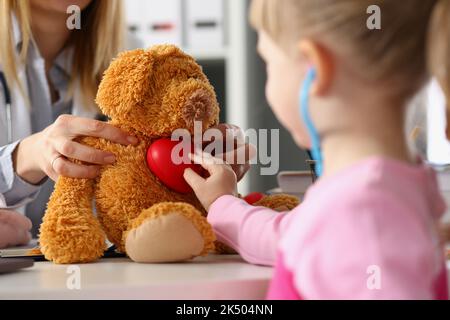 La bambina gioca con il giocattolo peluche su appuntamento del medico. Il bambino ascolta il giocattolo morbido con stetoscopio e tiene il cuore Foto Stock