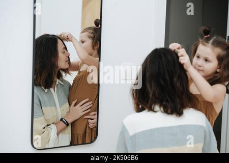 Carina bambina spazzolando i capelli della madre di fronte ad uno specchio. La mamma la abbraccia in un impulso. Foto Stock