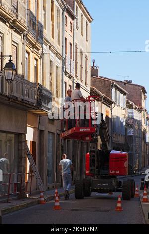 Limoux Aude France 19.4.22 due uomini gestiscono una piattaforma aerea rossa in una strada stretta di vecchi edifici con balconi in ferro. Un giovane su scooter elettrico Foto Stock