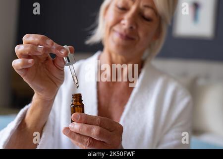 Felice donna caucasica anziana seduta sul letto in camera da letto, applicando siero Foto Stock