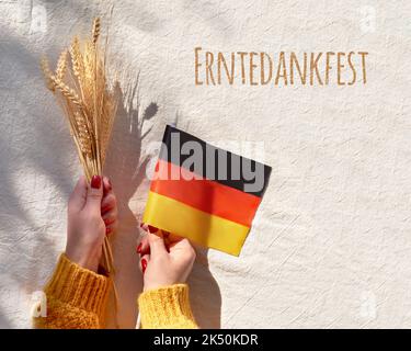 Happy Erntedankfest, Harvest Thanksgiving Day in lingua tedesca. Bandiera tedesca e mazzo di spighe di grano in mano su tessuto bianco beige. Giacitura piatta Foto Stock