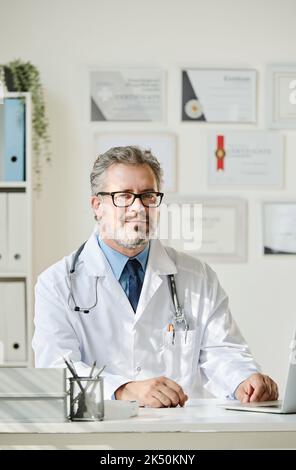 Ritratto di medico maturo in cappotto bianco seduto al suo posto di lavoro in ufficio Foto Stock