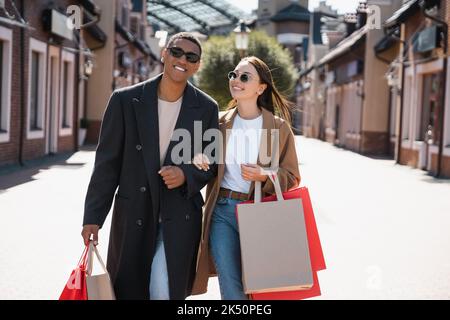 coppia interrazziale gioiosa in cappotti e occhiali da sole che trasportano i sacchetti di shopping sulla strada della città, immagine di riserva Foto Stock