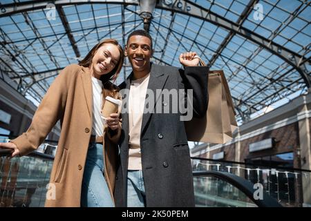 coppia interrazziale gioiosa in cappotti alla moda in possesso di borse da shopping e caffè per andare, mentre sorridendo alla macchina fotografica sotto tetto di vetro, immagine stock Foto Stock