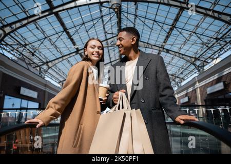 donna allegra con caffè per andare a guardare la macchina fotografica vicino all'uomo afroamericano con le borse di shopping, immagine di scorta Foto Stock