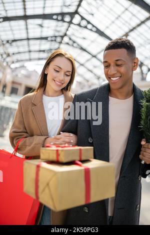 gioiosa coppia multietnica in cappotti alla moda guardando sfocata scatole regalo, immagine stock Foto Stock