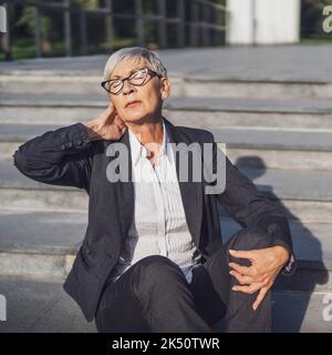 Donna d'affari anziana seduta davanti all'edificio dell'azienda. È stanca e ha dolore al collo. Foto Stock