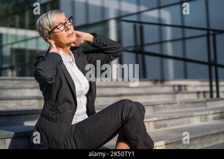 Donna d'affari anziana seduta davanti all'edificio dell'azienda. È stanca e ha dolore al collo. Foto Stock