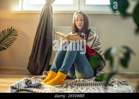 Una donna avvolta in un plaid è seduta sul pavimento con il tè caldo e si scalda vicino ad un radiatore caldo. Il concetto di economia e la crisi di ene Foto Stock
