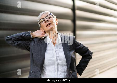 La donna d'affari anziana sta avendo sprain nel collo. Si trova di fronte alla costruzione dell'azienda. Foto Stock