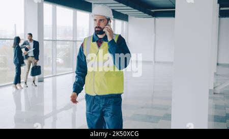 ritratto di ingegnere indossare casco e giubbotto parlare sul telefono cellulare in un nuovo edificio a piedi in una sala vuota concentrato sulla conversazione Foto Stock