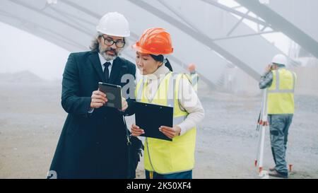 Giovane ingegnere donna asiatica sta parlando con uomo maturo caucasico investitore discutere progetto di costruzione leggere carte e utilizzare tablet all'aperto Foto Stock