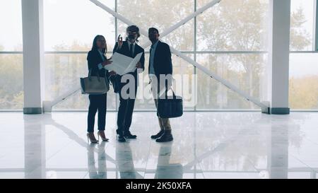 Gli investitori discutono di nuovo edificio industriale studiando il progetto in piedi contro le grandi finestre panoramiche in una stanza vuota che parla e che scuote le mani Foto Stock