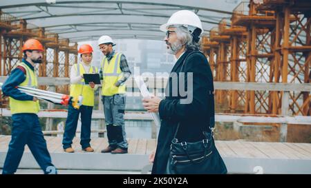 L'investitore maturo sicuro dell'uomo sta lavorando nel modello holding del luogo di costruzione mentre i lavoratori sono occupati in background. Concetto di persone e affari. Foto Stock