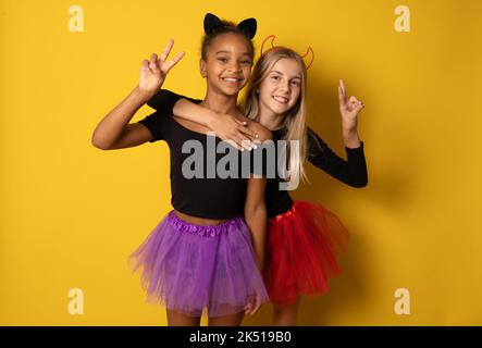 Immagine di ragazze multinazionali ridenti in costumi neri e colorati di Halloween isolati sopra la parete gialla Foto Stock