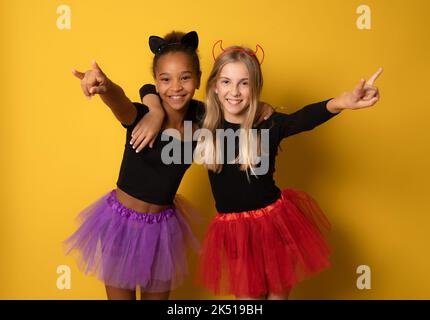 Immagine di ragazze multinazionali ridenti in costumi neri e colorati di Halloween isolati sopra la parete gialla Foto Stock