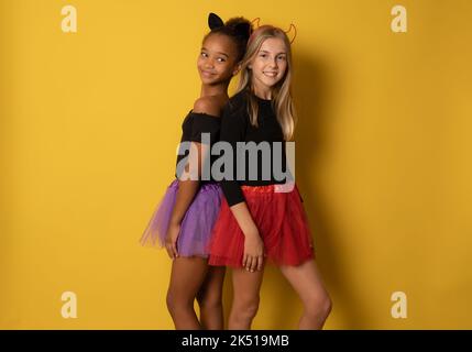 Immagine di ragazze multinazionali ridenti in costumi neri e colorati di Halloween isolati sopra la parete gialla Foto Stock