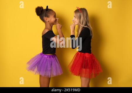 Immagine di ragazze multinazionali ridenti in costumi neri e colorati di Halloween isolati sopra la parete gialla Foto Stock