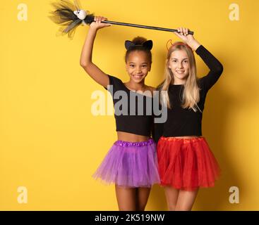 Immagine di ragazze multinazionali ridenti in costumi neri e colorati di Halloween isolati sopra la parete gialla Foto Stock