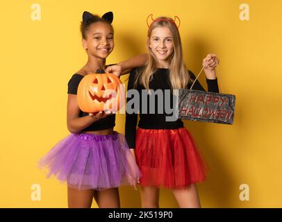 Immagine di ragazze multinazionali ridenti in costumi neri di Halloween con zucche isolate su sfondo giallo. Foto Stock