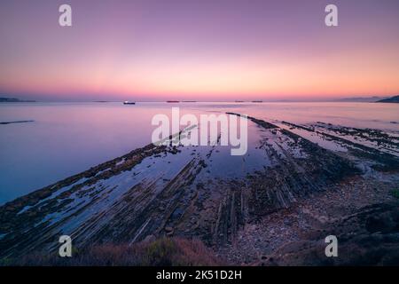 Acque tranquille di mare che lavano rocce sulla costa sotto il cielo viola tramonto in serata con navi a vela a distanza Foto Stock