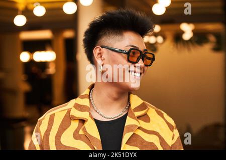 Sorridente giovane non binary maschio in camicia elegante con occhiali da sole e accessori con imperfezioni della pelle del viso che guarda lontano Foto Stock