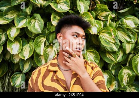 Giovane filippino da sogno, giovane e premuroso, non binary, che indossa una camicia fresca con orecchini in piedi nel giardino verde che guarda lontano Foto Stock