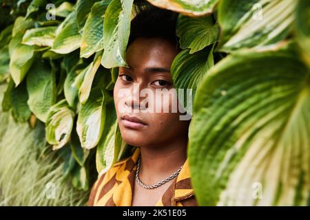 Giovane filippino da sogno, giovane e premuroso, non binary, che indossa una camicia fredda con orecchini in piedi nel giardino verde guardando la macchina fotografica Foto Stock
