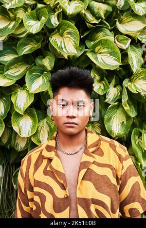 Giovane filippino da sogno, giovane e premuroso, non binary, che indossa una camicia fredda con orecchini in piedi nel giardino verde guardando la macchina fotografica Foto Stock
