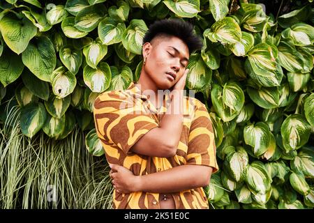Giovane filippino da sogno non binary uomo che indossa una camicia cool con orecchini che si abbracciano con amore in piedi con gli occhi chiusi in giardino verde Foto Stock