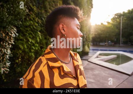 Giovane filippino da sogno non binary uomo che indossa una camicia fredda con orecchini in piedi con gli occhi chiusi nel giardino verde Foto Stock