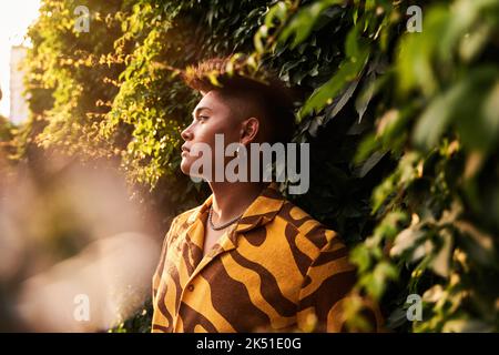 Giovane filippino da sogno, giovane e premuroso, non binary, che indossa una camicia fresca con orecchini in piedi nel giardino verde che guarda lontano Foto Stock