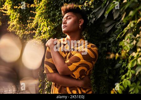 Giovane filippino da sogno non binary uomo che indossa una camicia cool con orecchini che si abbracciano con amore in piedi con gli occhi chiusi in giardino verde Foto Stock