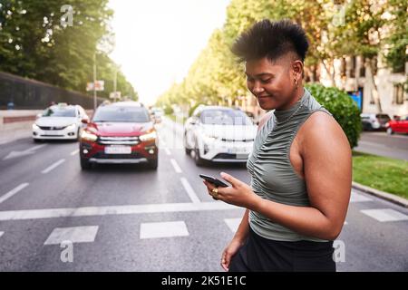 Donna transessuale filippino sorridente in alto e gonna messaggio di controllo sullo smartphone, mentre si attraversa la strada vicino al parco in città Foto Stock