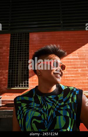 Da sotto felice filippino non binary persona in abiti eleganti e occhiali da sole guardando lontano e sorridendo contro muro di mattoni illuminato dal sole Foto Stock