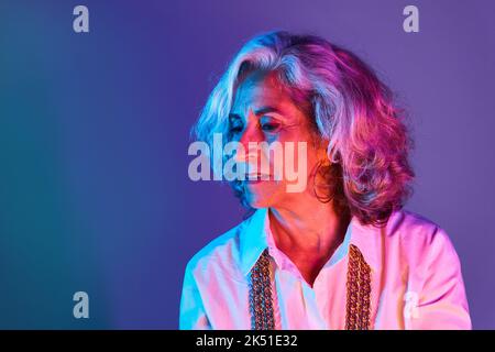 Ritratto di donna anziana fiduciosa con capelli corti in argento e in camicia bianca guardando verso il basso in luci rosa e blu Foto Stock