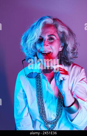 Donna anziana felice in camicia bianca con elegante collana guardando la fotocamera con sorriso e mordente occhiali da sole sotto colorata luce al neon Foto Stock