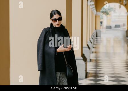 Seria giovane femmina etnica con capelli scuri in elegante cappotto nero e occhiali da sole appoggiati sulla colonna e guardando la macchina fotografica mentre si è in piedi in passa arcuata Foto Stock