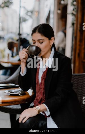 Imprenditrice etnica adulta autoassicurata con capelli scuri in elegante camicia bianca e cappotto nero bere una tazza di caffè in Street cafè Foto Stock