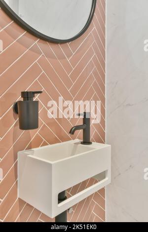 Lavabo in ceramica con rubinetti e dispenser con sapone liquido collocato su una parete piastrellata rosa e specchio in bagno bianco a casa Foto Stock