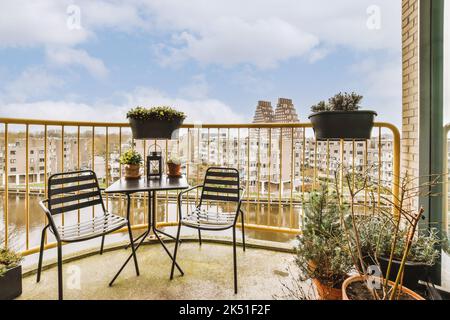 Sedie al tavolo nero posto su ampio balcone di casa con recinzione in metallo e fiori in vaso contro la costruzione il giorno d'estate Foto Stock