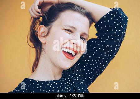 Felice giovane auto assicurare femmina con le labbra rosse in elegante vestito punteggiato sorridendo luminoso e winking alla macchina fotografica mentre tocca lunghi capelli scuri contro Foto Stock