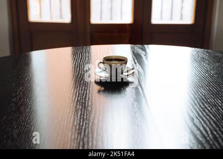 Tazza di espresso nero o caffè americano su un grande tavolo di legno sotto i raggi del sole del mattino. Vapore delicato su bevanda. Cucchiaio piccolo sul piatto. Copia spazio fo Foto Stock