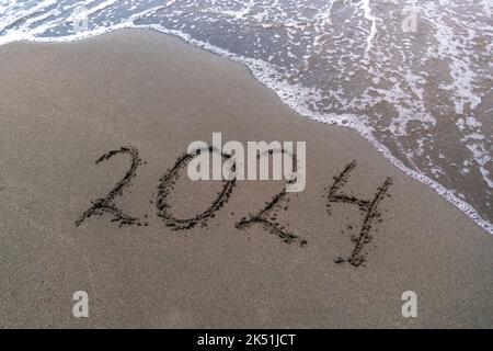 Testo felice anno nuovo 2024 su una spiaggia di sabbia con una piccola onda. Foto di sfondo astratta del prossimo anno 2023 Foto Stock