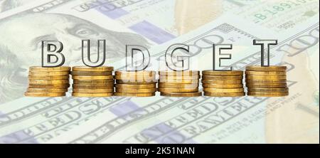 La parola 'Budget' fatta di lettere di vetro, in piedi su pile di monete sullo sfondo di banconote in dollari. Il concetto di un bilancio pulito. Foto Stock