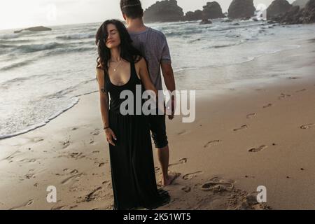 intera lunghezza di donna tatuata in vestito e uomo in piedi sulla spiaggia e tenendo le mani vicino oceano, immagine stock Foto Stock