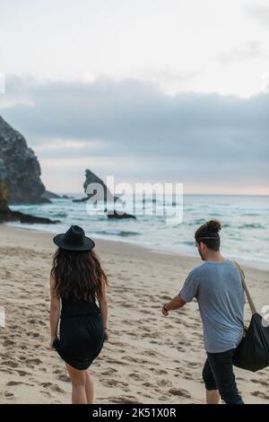 vista posteriore della donna tatuata e dell'uomo bearded con il sacchetto che cammina insieme sulla spiaggia sabbiosa, immagine di scorta Foto Stock