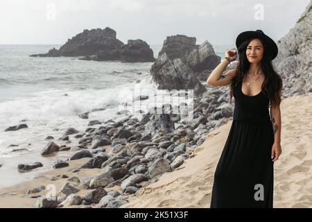 donna allegra e tatuata in abito nero e cappello in piedi sulla spiaggia di sabbia in portogallo, immagine stock Foto Stock