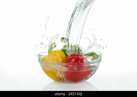 cascata che si riversa su una ciotola di vetro con peperoni multicolore con spruzzi su fondo bianco. Foto Stock