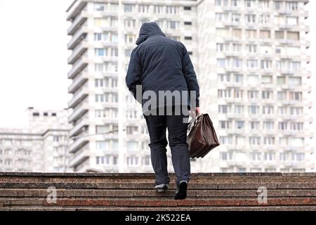 Uomo in giacca con cappuccio con valigetta in pelle che sale le scale in pietra in città durante la pioggia. Concetto di carriera, di successo, di spostamento verso l'alto Foto Stock